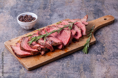 Slices of medium rare roast beef meat on wooden cutting board, pepper and rosemary twigs on gray marble background. Gourmet food. Raw meat beef steak.