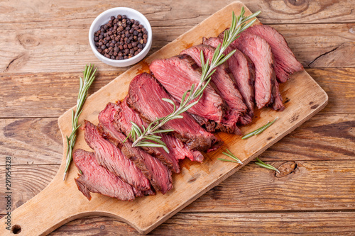 Slices of medium rare roast beef meat on wooden cutting board, pepper and rosemary branches on wooden background. Top view. Gourmet food. Raw meat beef steak.