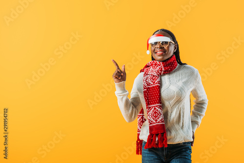 African American woman in winter sweater and Christmas glasses pointing with finger isolated on yellow