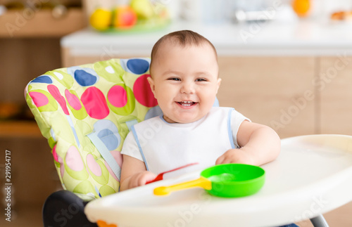 Cheerful infant sitting in high children chair toys in kitchen