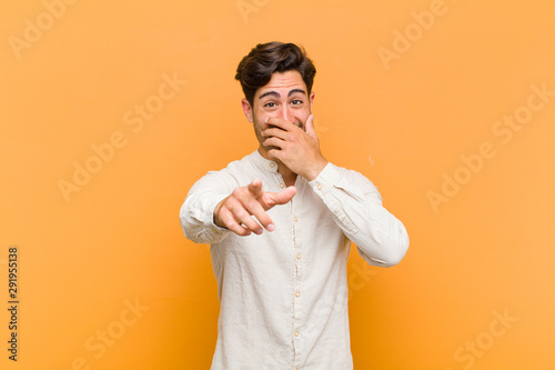 young handsome man laughing at you, pointing to camera and making fun of or mocking you against orange background