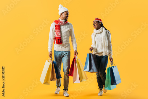 African American couple in winter outfit walking with shopping bags and looking at each other isolated on yellow