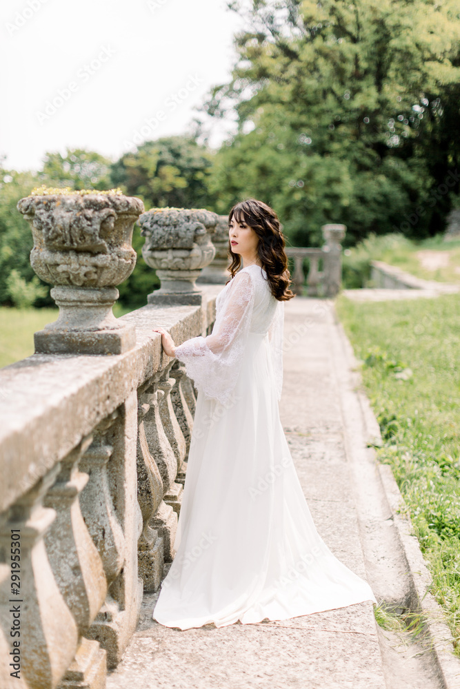 Amazing brunette Chinese Asian bride in luxury wedding dress, posing at old castle background, standing near the stone railings, wedding photo, European city, wedding day, portrait