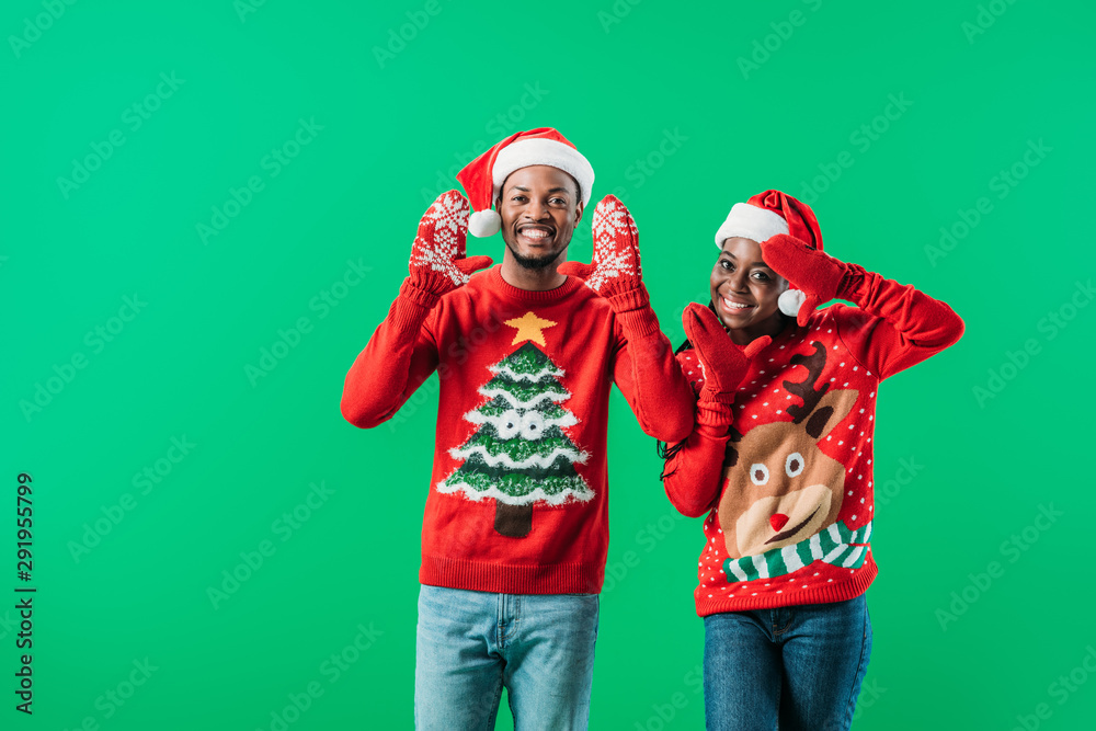 African American couple in red Christmas sweaters and Santa hats raising hands in winter gloves with pattern isolated on green