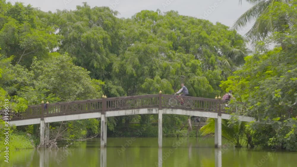 A daylight wide shot of a stilted cement bridge over the calm water of a pond with cyclists passing over it against the background of luscious trees in the forest.. A daylight wide shot of a stilted cement bridge over the calm water of a pond with cyclists passing over it against the background of luscious trees in the forest..