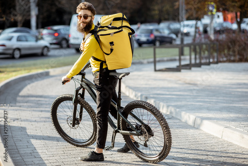Portrait of a young courier delivering food with a yellow thermal backpack on a bicycle in the city. Food delivery service concept
