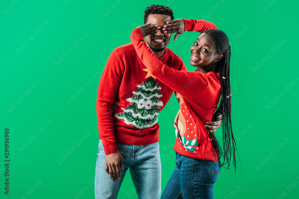 African American woman in Christmas sweater covering eyes with hands to man isolated on green African American woman in Christmas sweater covering eyes with hands to man isolated on green