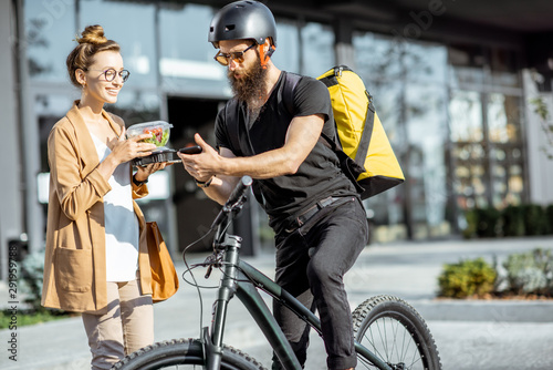 Happy businesswoman receiving fresh takaway lunches, standing near the office building with a male courier on the background. Takeaway restaurant food delivery concept