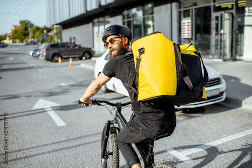 Young bearded courier delivering food with a yellow thermal backpack, riding a bicycle in the city. Food delivery service concept photo