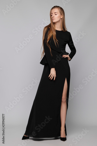 Full shot of young blonde European lady with long straight hair wearing a stylish black maxi dress decorated with an asymmetric frill, a basque and a side midthigh hem tear, girl's hand is on her hip.