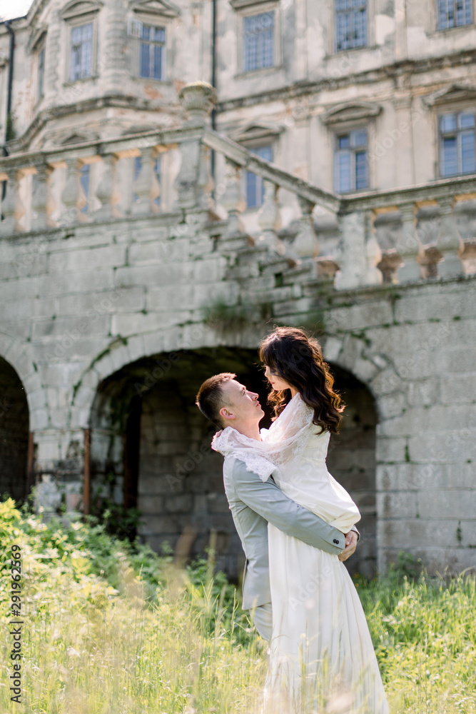 beautiful gorgeous Chinese bride and groom walking in sunny day and kissing. happy wedding couple hugging, man holds woman, outdoors near old castle. happy romantic moments