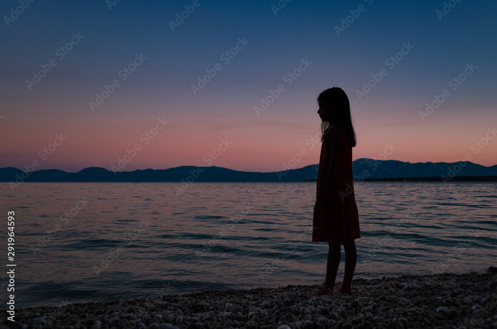 Fototapeta premium Silhouetter of a young girl standing on a beach looking on horizon after sunset