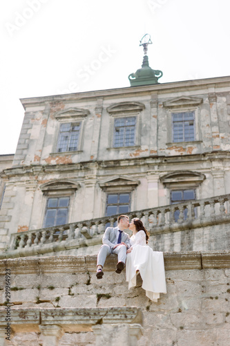 Beautiful Asian wedding couple poses while sitting on the stone wall of an old ruined castle