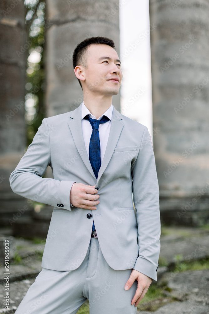 Elegant smiling handsome Asian groom in gray suit posing on the background of ancient stone columns in old castle Elegant smiling handsome Asian groom in gray suit posing on the background of ancient stone columns in old castle