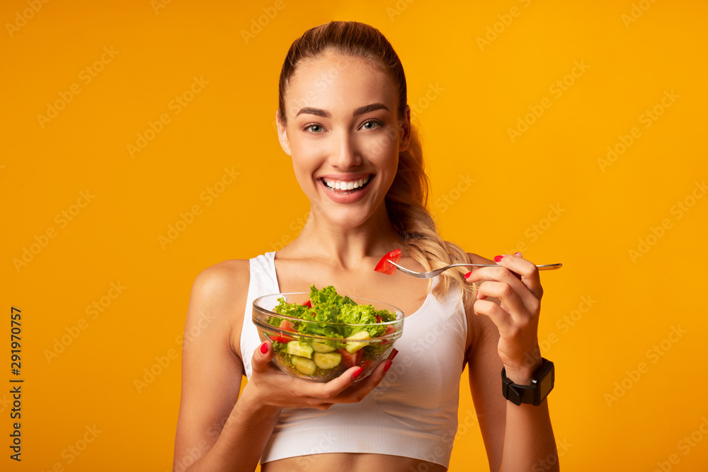 Fitness Girl Eating Vegetable Salad Standing Over Yellow Background - obrazy, fototapety, plakaty 