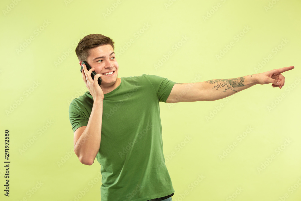 Caucasian young man's half-length portrait on green studio background. Beautiful male model in shirt. Concept of human emotions, facial expression, sales, ad. Talking on phone and pointing, smiling.