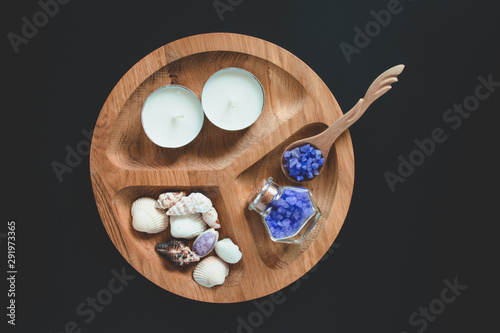 Seshels, bottle, lavender salts and spoon, white candle on the wooden round form. Black background.