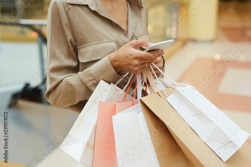 Close-up of young woman typing a message on her mobile phone and holding shopping bags in the store