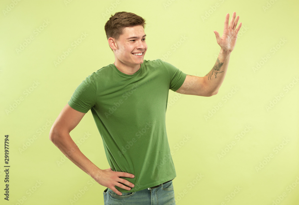 Caucasian young man's half-length portrait on green studio background. Beautiful male model in shirt. Concept of human emotions, facial expression, sales, ad. Greeting somebody and smiling. Caucasian young man's half-length portrait on green studio background. Beautiful male model in shirt. Concept of human emotions, facial expression, sales, ad. Greeting somebody and smiling.