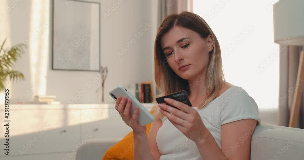 Close up of the young charming woman tapping on the smartphone while shopping online and entering credit card number in the cozy room, then turning her face and smiling to the camera. Portrait. Close up of the young charming woman tapping on the smartphone while shopping online and entering credit card number in the cozy room, then turning her face and smiling to the camera. Portrait.