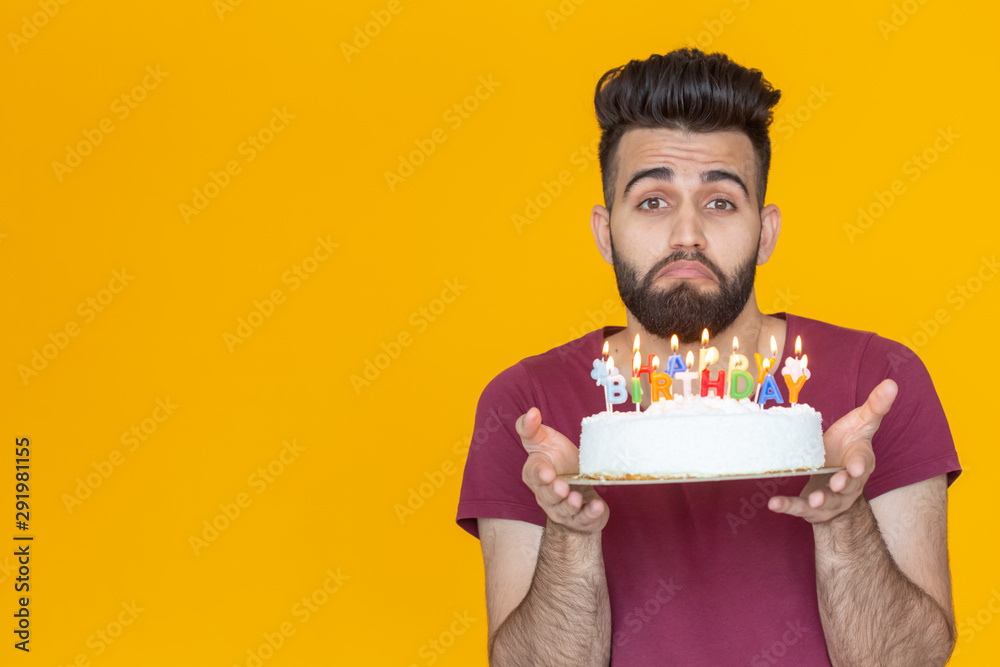 Crazy cheerful young guy holding a burning candle in his hands and a congratulatory homemade cake on a yellow background. Birthday and anniversary celebration concept. Crazy cheerful young guy holding a burning candle in his hands and a congratulatory homemade cake on a yellow background. Birthday and anniversary celebration concept.