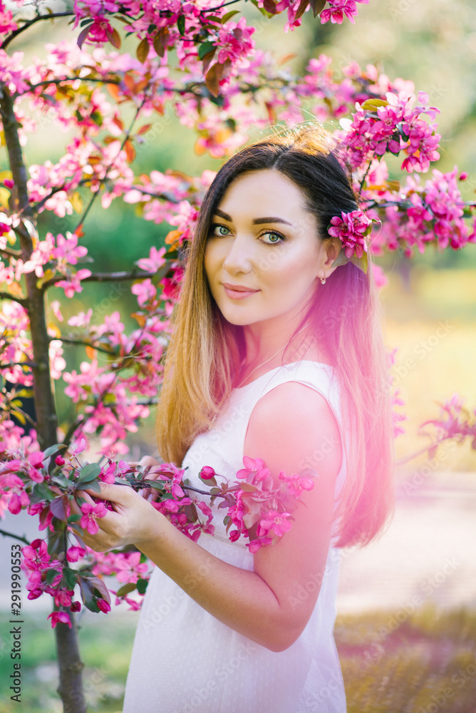 Fototapeta premium A beautiful girl with long straight hair and big eyes stands in the flowers of a pink Apple tree. Professional makeup