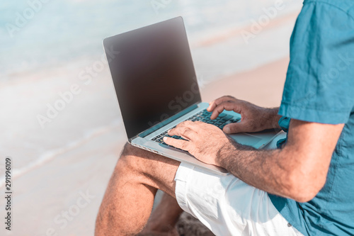 Adult male of mediterranean race using laptop while sitting on sandy beach. Freedom and travel concept
