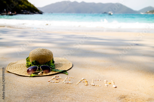 Hat and sunglasses over beach sand. Vacation concept. Beach written on sand with shells.