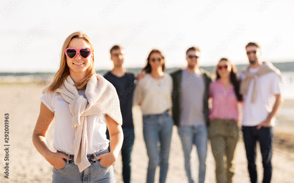 Obraz premium friendship, valentine's day and people concept - happy woman in heart-shaped sunglasses with group of friends on beach in summer