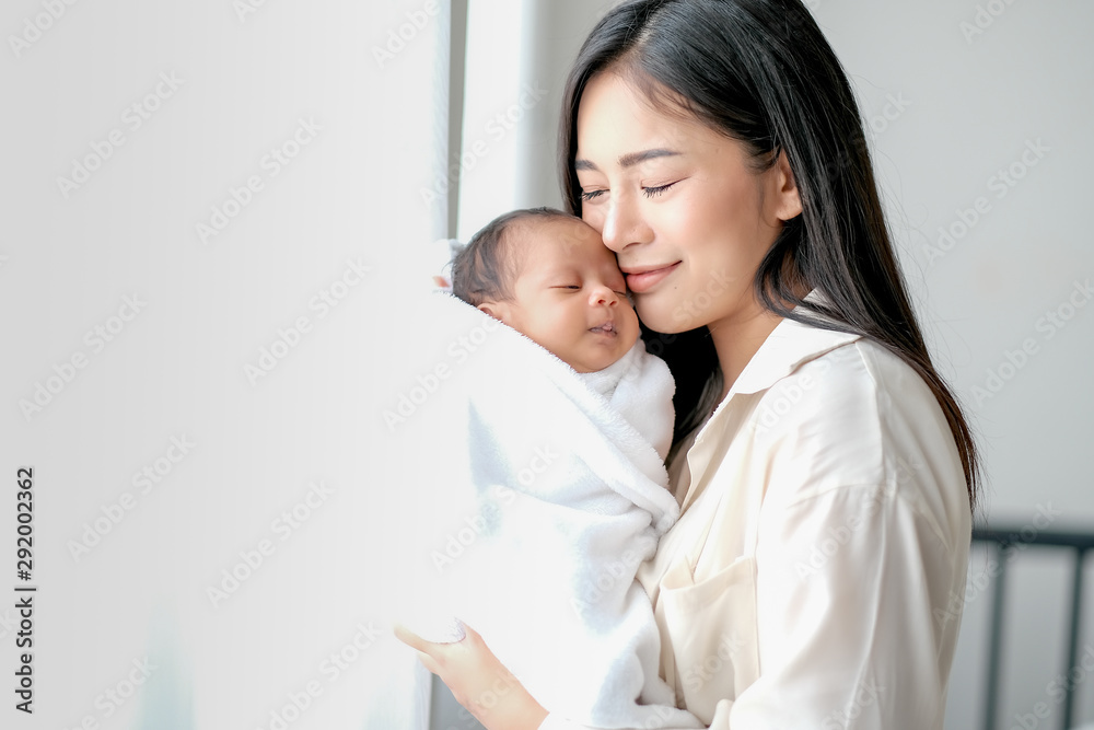 White shirt Asian mother is kissing her newborn baby in bedroom in front of glass windows with white curtain to show love and family bonding. White shirt Asian mother is kissing her newborn baby in bedroom in front of glass windows with white curtain to show love and family bonding.