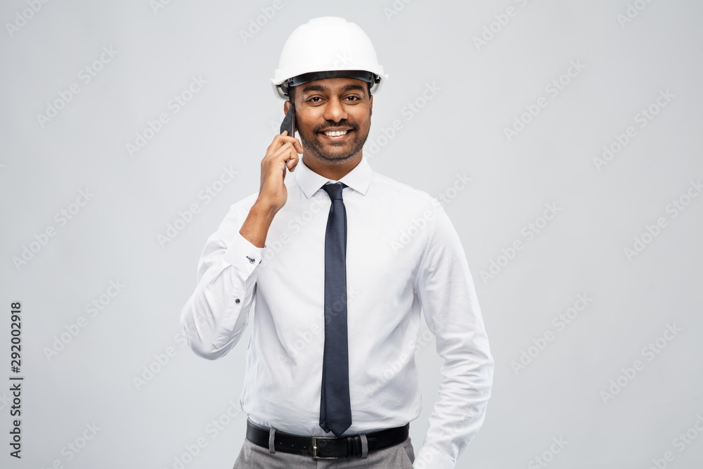 architecture, construction business and people concept - smiling indian male architect in helmet calling on smartphone over grey background architecture, construction business and people concept - smiling indian male architect in helmet calling on smartphone over grey background