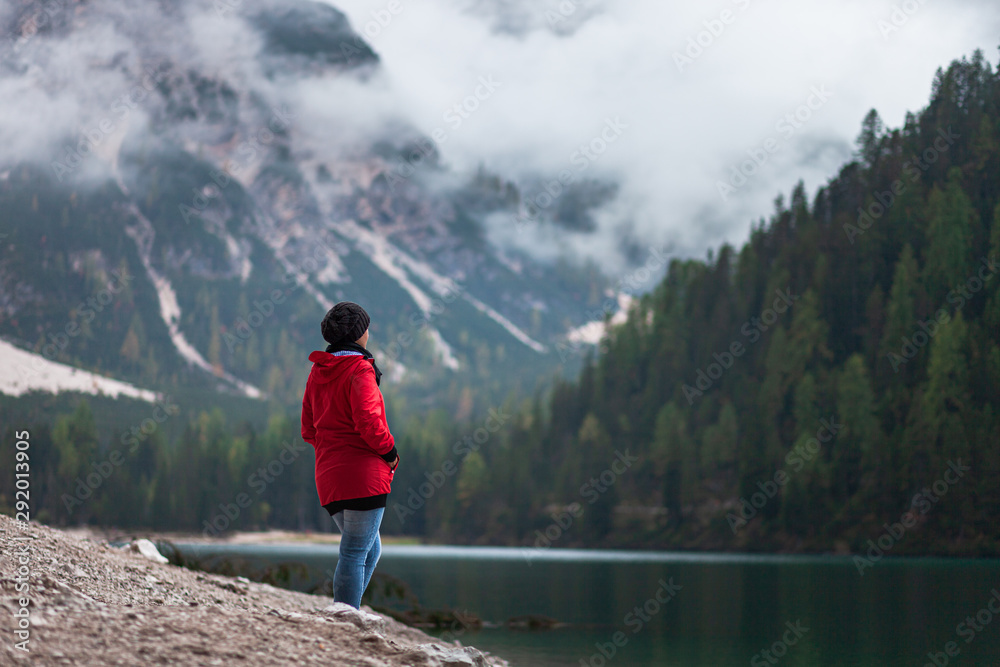 Women with red rain jacket enjoying the peaceful mystic view of a mountain lake with cloudy dramatic mountains in the background on a rainy autumn day.