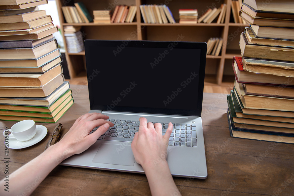 Hands are on the laptop in the library on the table with many books and a cup of coffee