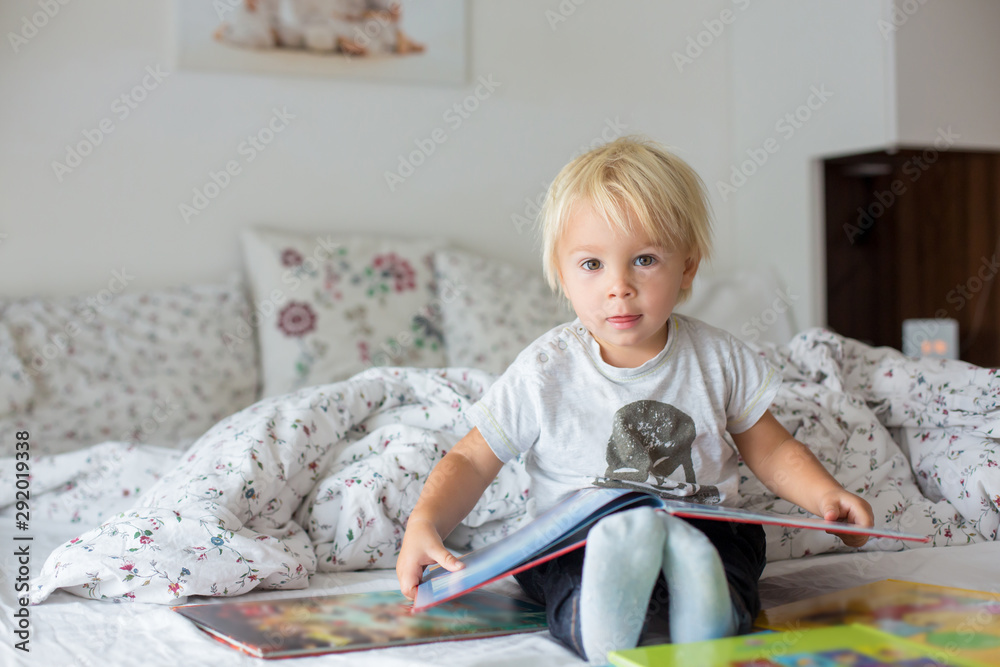 Sweet toddler boy, reading book at home, sitting in bed, lots of books Sweet toddler boy, reading book at home, sitting in bed, lots of books