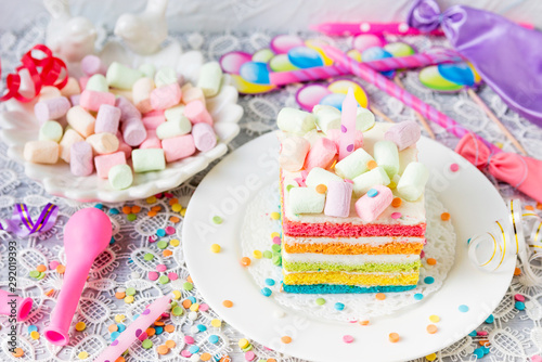 Rainbow birthday cake with marshmallows. Balls, candles, confetti on the festive table.