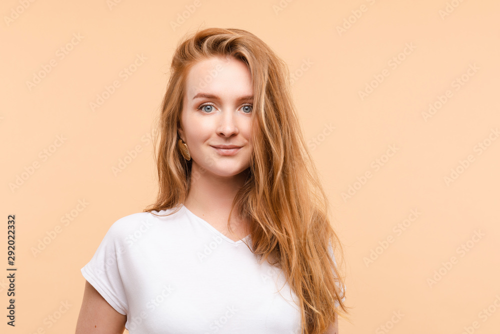 Studio portrait of lovely beautiful young adult woman with blue eyes and long wavy red hair in white t-shirt looking at camera. Isolate on pale yellow background. Studio portrait of lovely beautiful young adult woman with blue eyes and long wavy red hair in white t-shirt looking at camera. Isolate on pale yellow background.