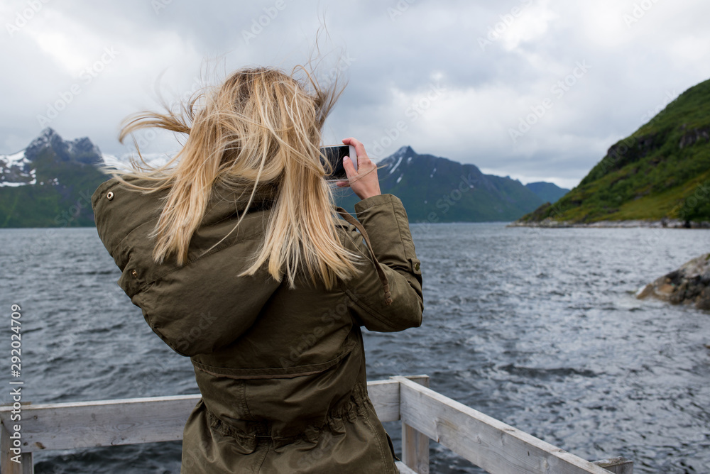 Fototapeta premium Blond hair woman walking along the pier enjoying scenic views. Ocean and rocks landscape. Lonely girl. Travel, adventure. Sense of freedom, lifestyle. Explore North Norway. Summer in Scandinavia