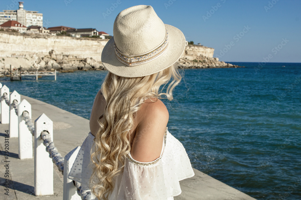 Beautiful blonde girl, with long hair in a hat at the sea, stands with ...