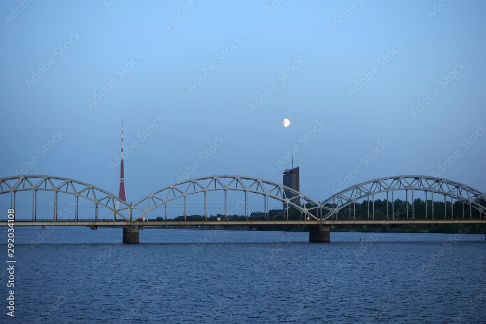 Long metal railroad arched bridge across the Daugava river in Riga against stand alone building, TV tower and bright moon forward view on sunsetsky Long metal railroad arched bridge across the Daugava river in Riga against stand alone building, TV tower and bright moon forward view on sunsetsky