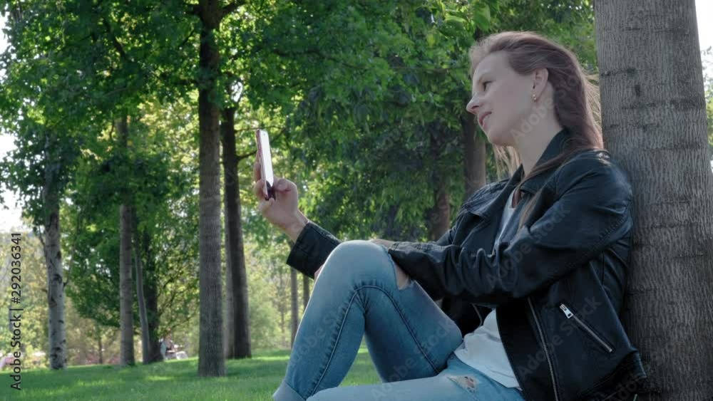 Young beautiful girl in street clothes in a park. Holding a smartphone in his hands, sitting on the grass under a tree. He takes selfies for social networks, looking for the best angle and pose. Young beautiful girl in street clothes in a park. Holding a smartphone in his hands, sitting on the grass under a tree. He takes selfies for social networks, looking for the best angle and pose.