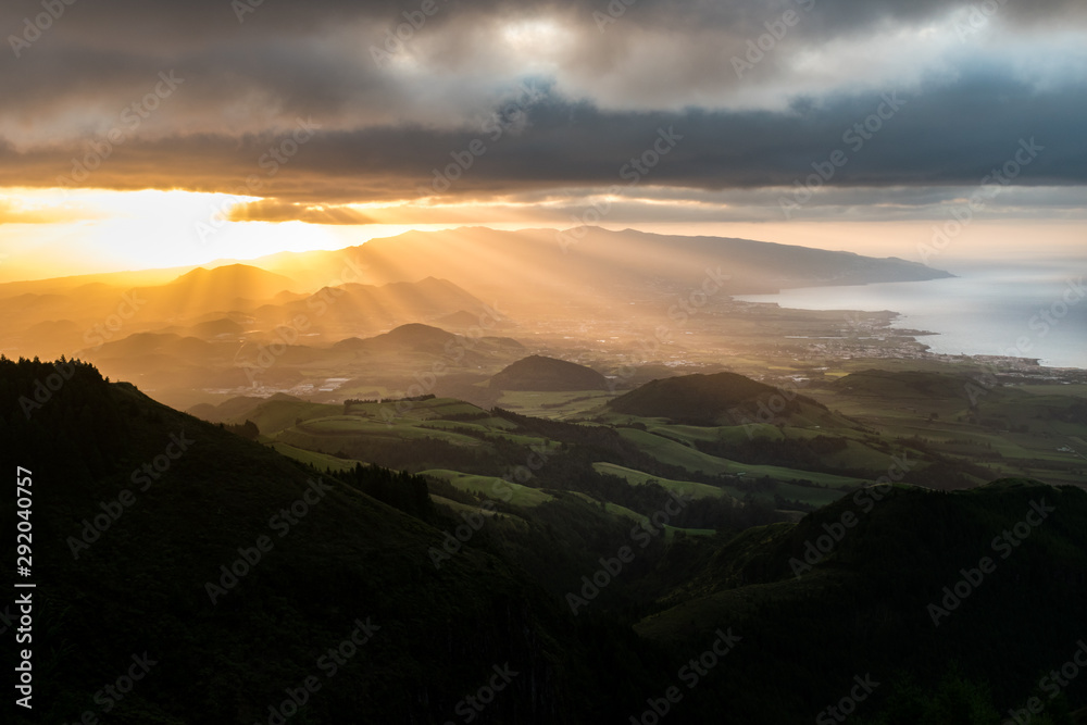 Stunning sunset view of the volcanic landscape on the island of São Miguel in the Azores archipelago. Volcanic cones are illuminated by sun rays under a dramatic cloudy sky with the Atlantic ocean.