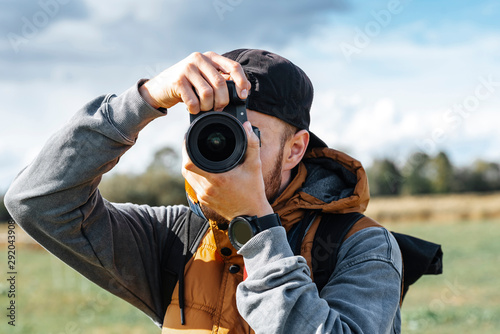 Photographer in brown vest and black hat taking pictures with his camera during sunny summer day. 