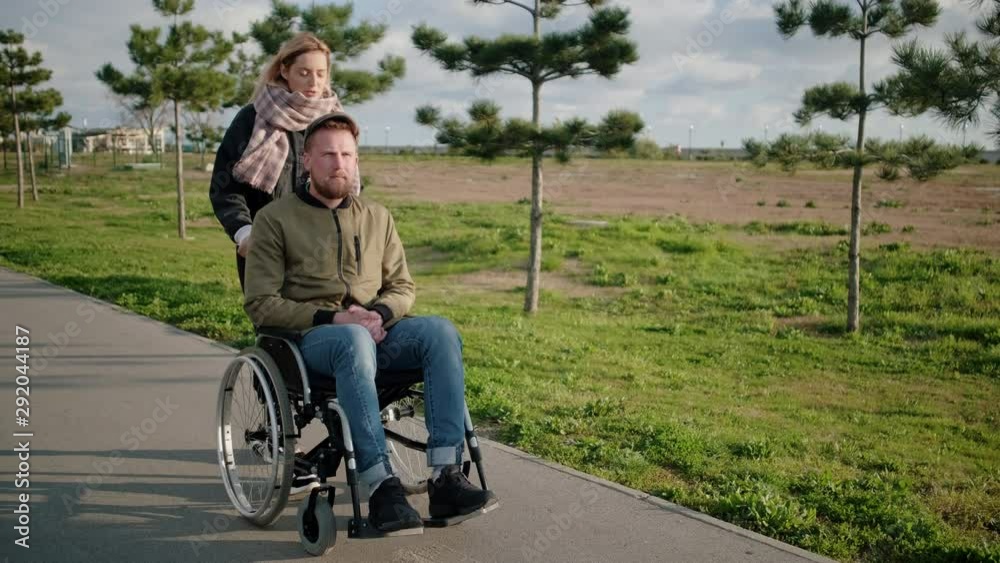 Man is sitting in invalid carriage and his wife is helping him, pushing it Man is sitting in invalid carriage and his wife is helping him, pushing it