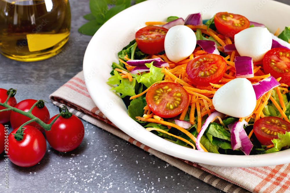 close up green leafy salad with carrot, tomato, onion and cheese near tomatoes on gray table with olive oil and basil leaves in the background close up green leafy salad with carrot, tomato, onion and cheese near tomatoes on gray table with olive oil and basil leaves in the background