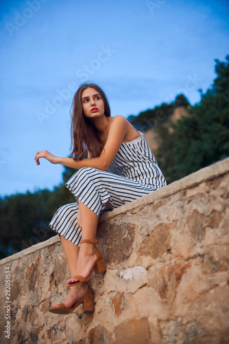 Beautiful woman wearing striped overalls and stylish sandals sitting on a stone wall .