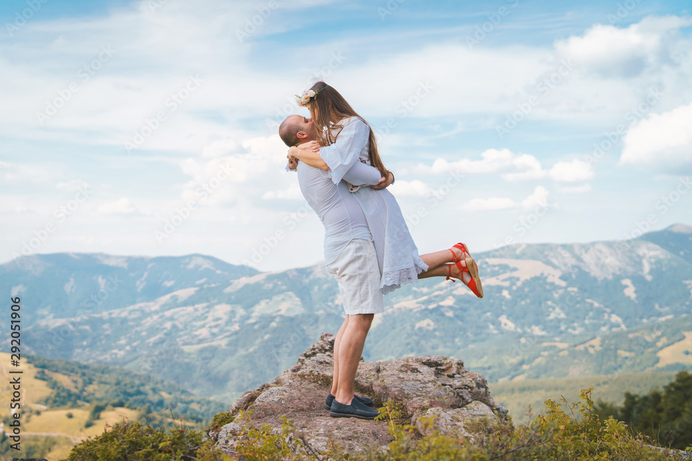 Fototapeta premium Young couple wearing white on the top of the mountain rock man holding his girl friend wife fiancee up hugging and kissing couple in love