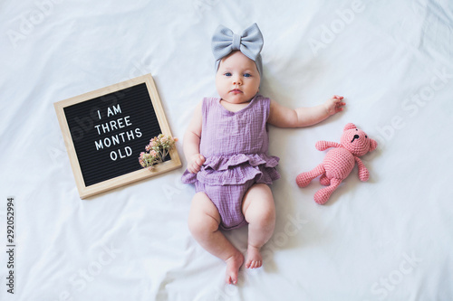 Three months old baby girl laying down on white background with letter board and teddy bear.