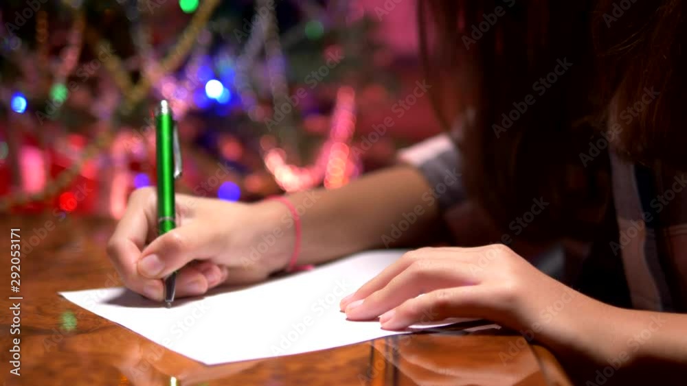 teen girl sits at a table near a decorated Christmas tree and writes a Christmas letter to Santa Claus teen girl sits at a table near a decorated Christmas tree and writes a Christmas letter to Santa Claus
