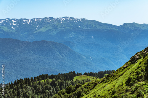 Green mountain ridges, surrounded by high mountains. Snow-capped mountain peaks on the horizon. Krasnaya Polyana, Sochi, Caucasus, Russia.
