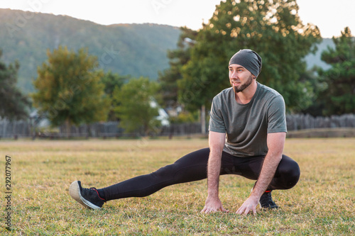 Sports and yoga. A man with a beard, in sportswear smiling and performs stretching on the grass before training